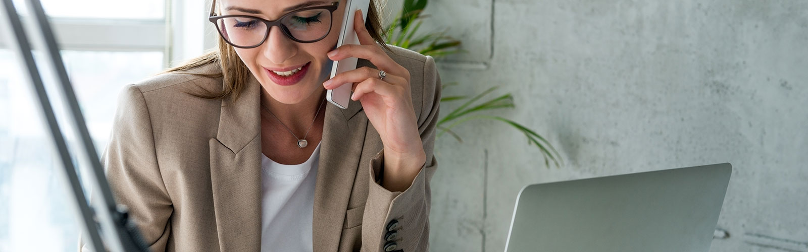 A woman in a work environment wearing a blazer and glasses, looking down as she talks on her smartphone