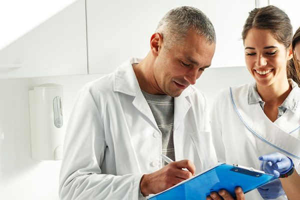 Implant dentist in lab coat holding a blue clipboard while consulting with two colleagues in dental office lab