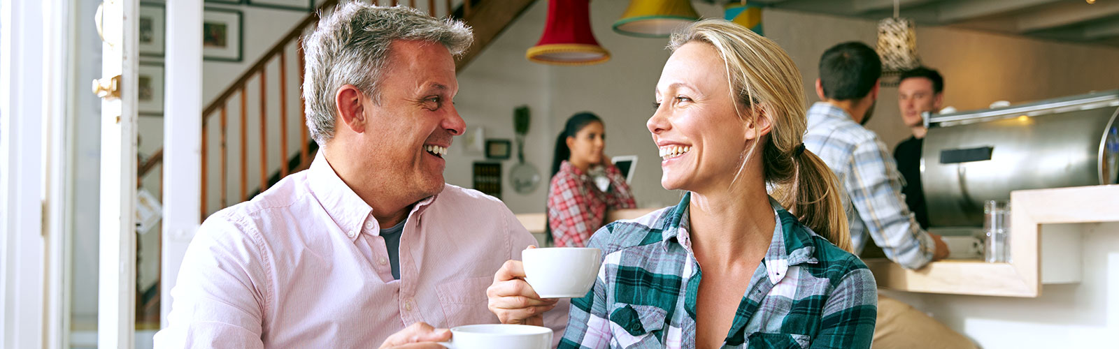 A man and woman in a coffee shop, sitting down and looking at each other while smiling and “cheers”-ing their coffee mugs